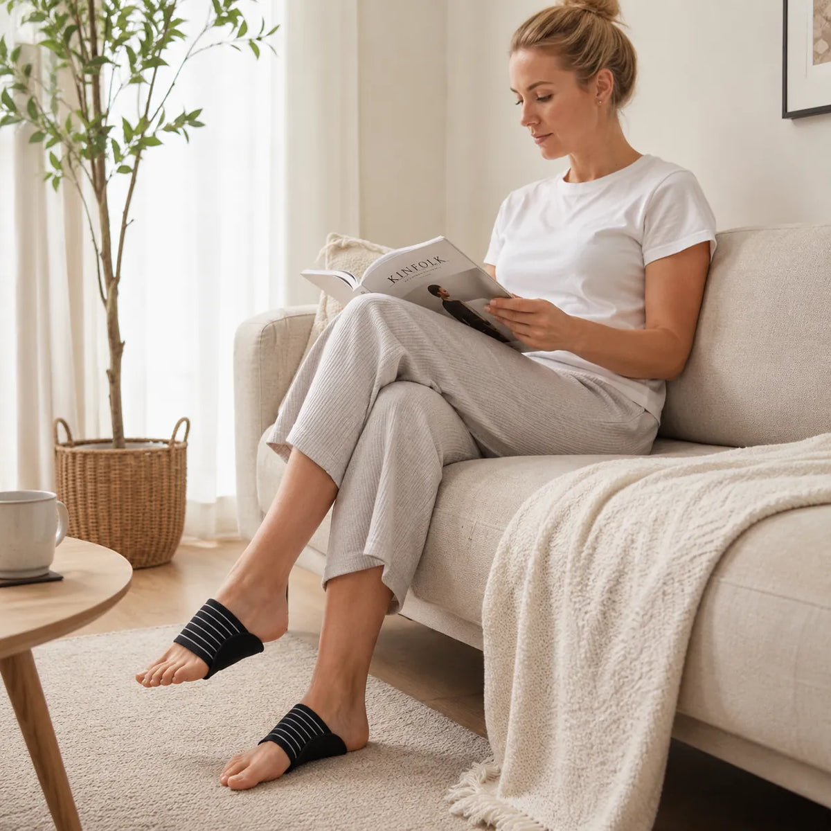 Une femme est assise sur un canapé et lit en portant des soutiens ergonomiques de voûte plantaire à sangle élastique.